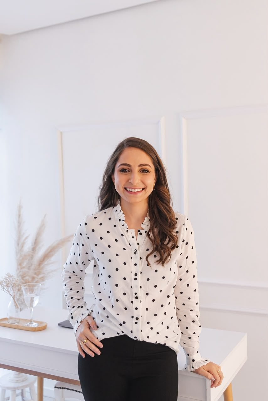 Dra. Cristina Haygert sorrindo, de camisa de bolinhas, apoiada na mesa do consultório